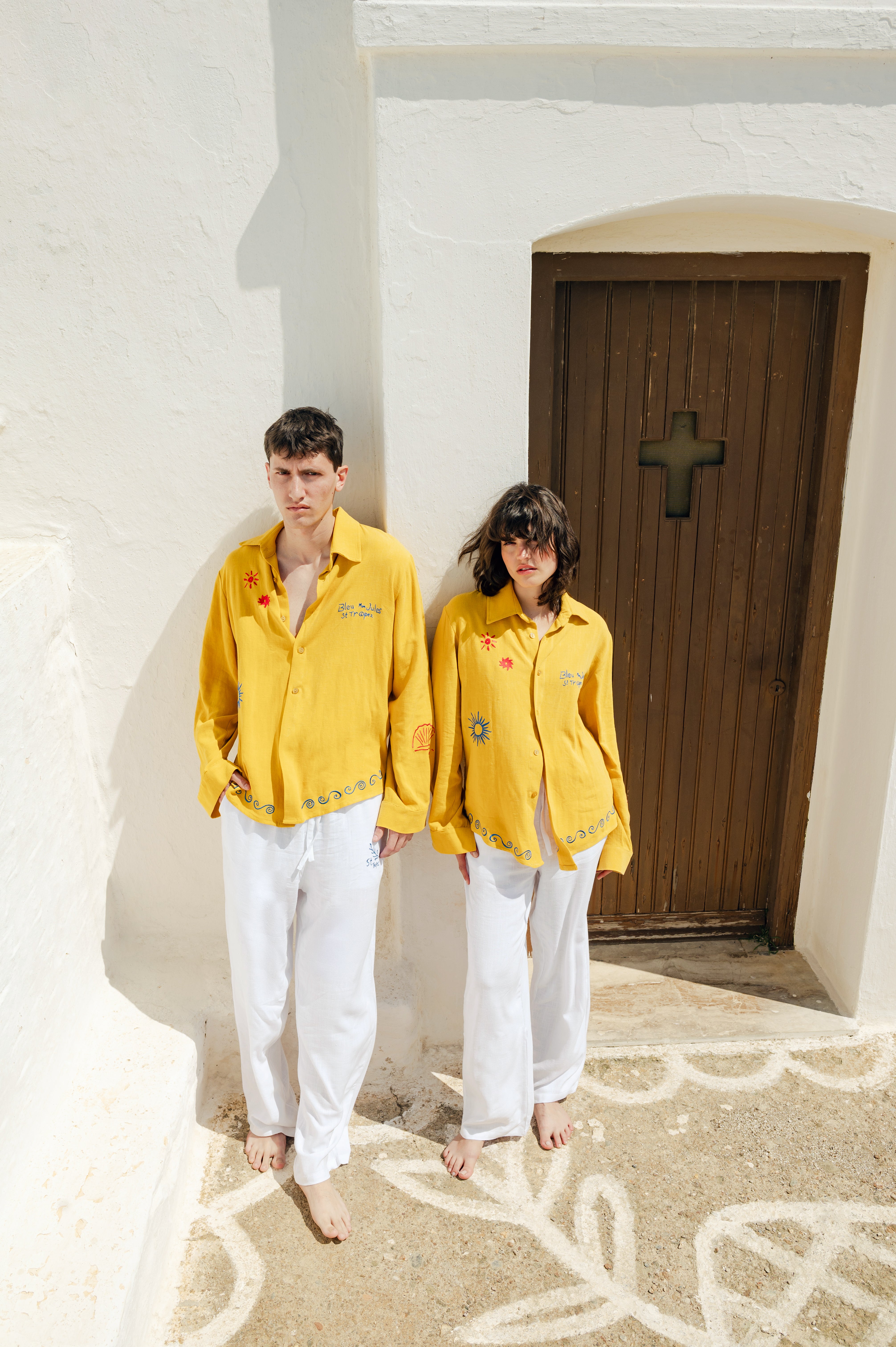 Couple en look d’été devant une porte grecque Duo vêtu de blanc et de jaune, ambiance Mykonos, lumière dorée et simplicité élégante.