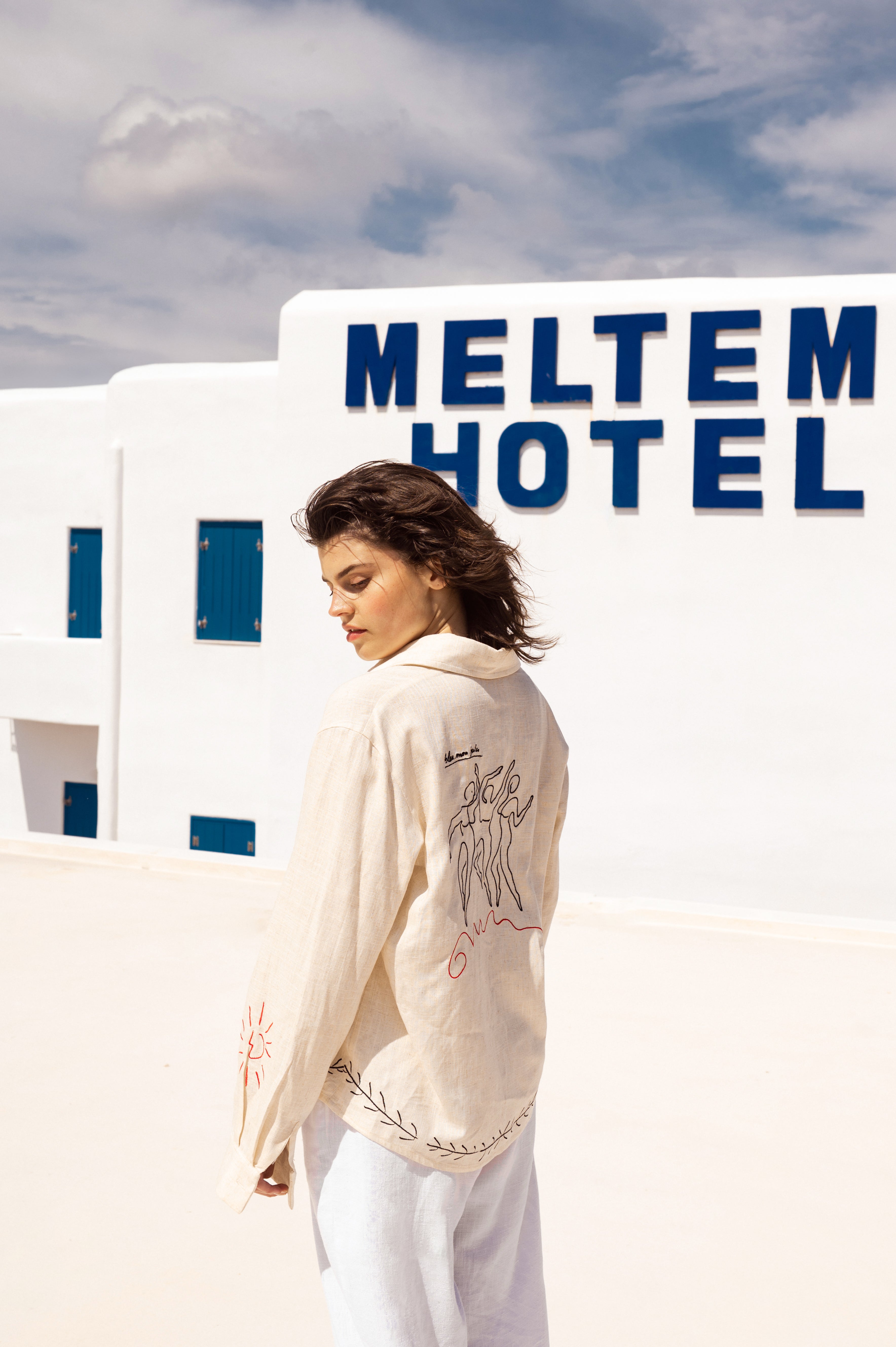 Femme en chemise jaune devant l’hôtel Meltem Look éclatant face à la mer Égée, chemise jaune et lumière blanche, esprit Riviera grecque.