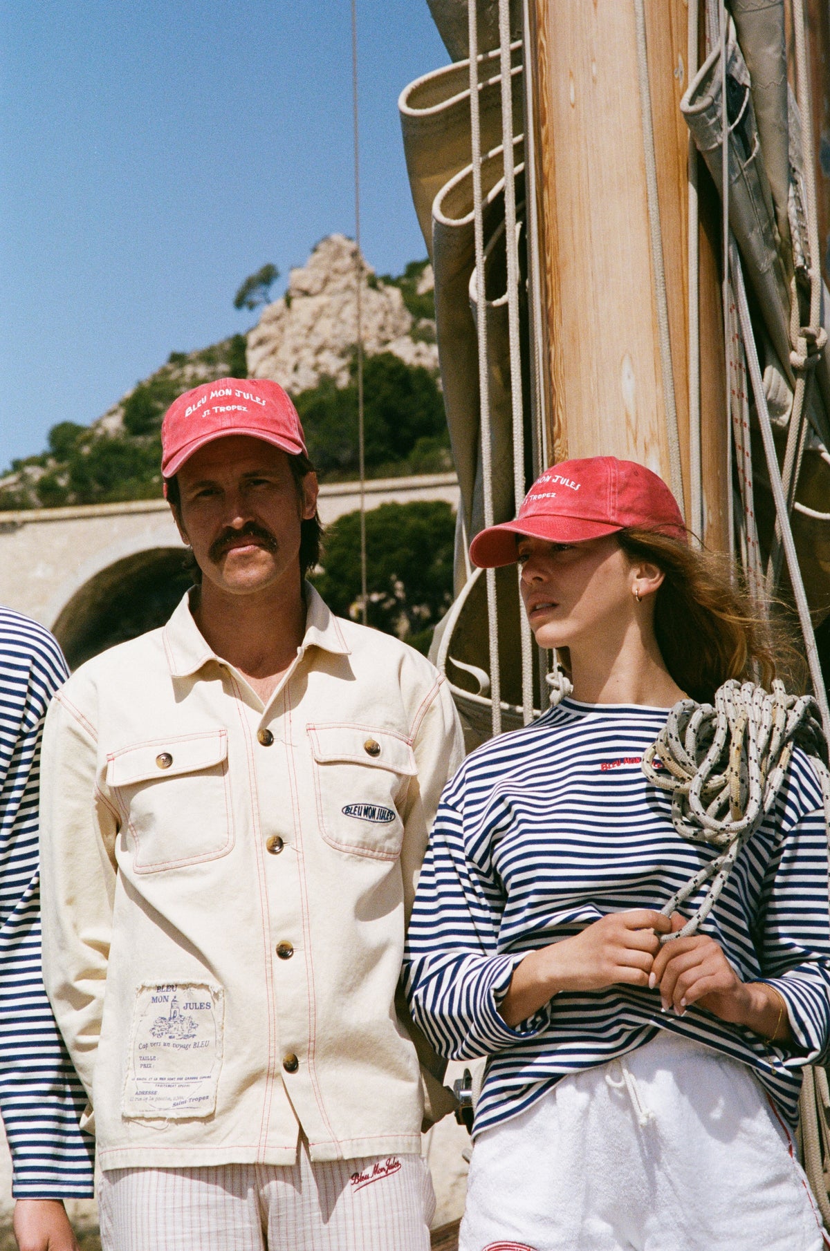 Couple portant casquette rouge Bleu mon Jules brodée, chemise blanche et rayée nautique devant bateau en Méditerranée