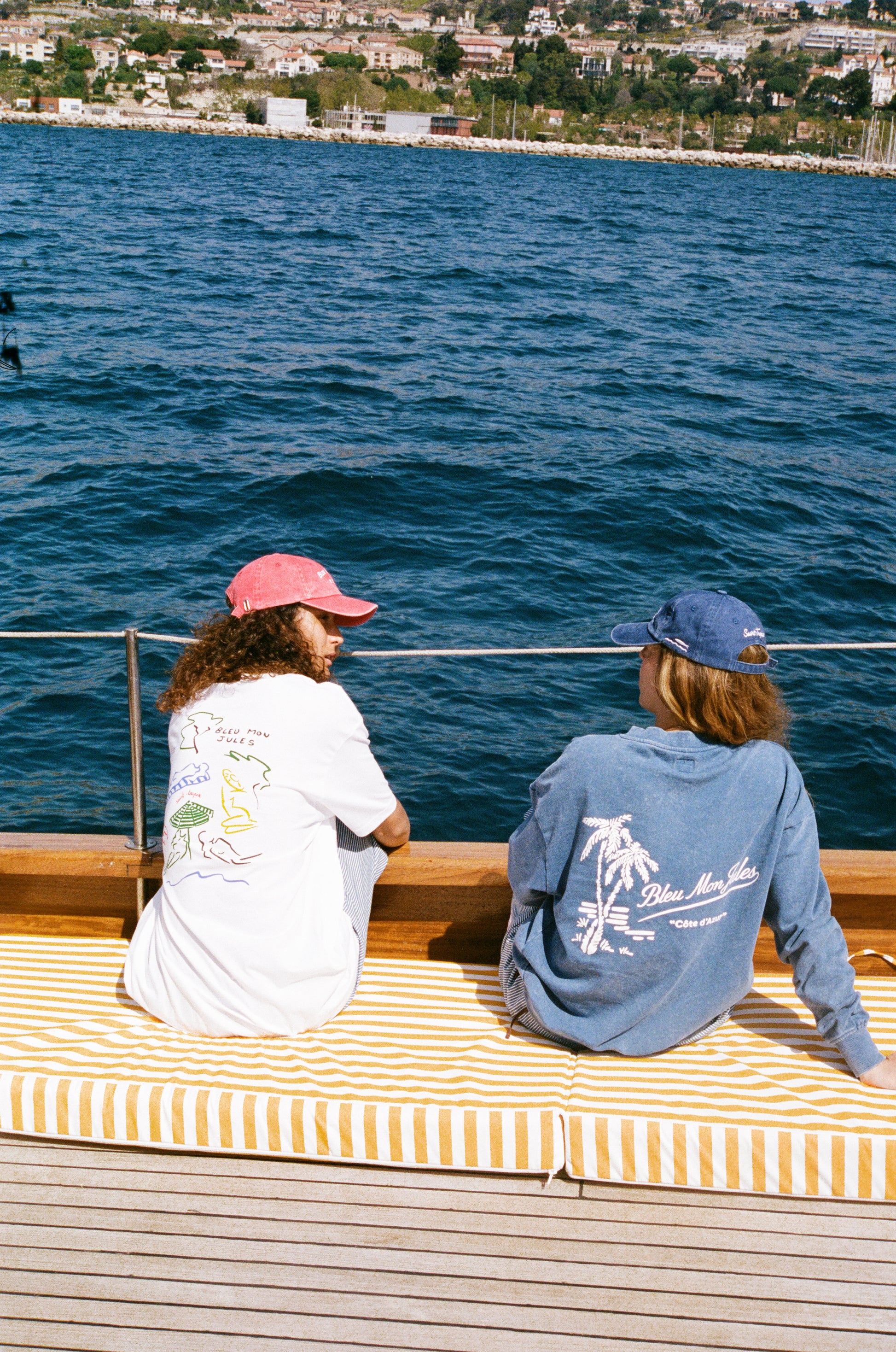 Deux personnes assises dos à la mer portant tee-shirts Bleu mon Jules, l'un blanc avec dessin Saint-Tropez style Miró, l'autre bleu marine avec logo