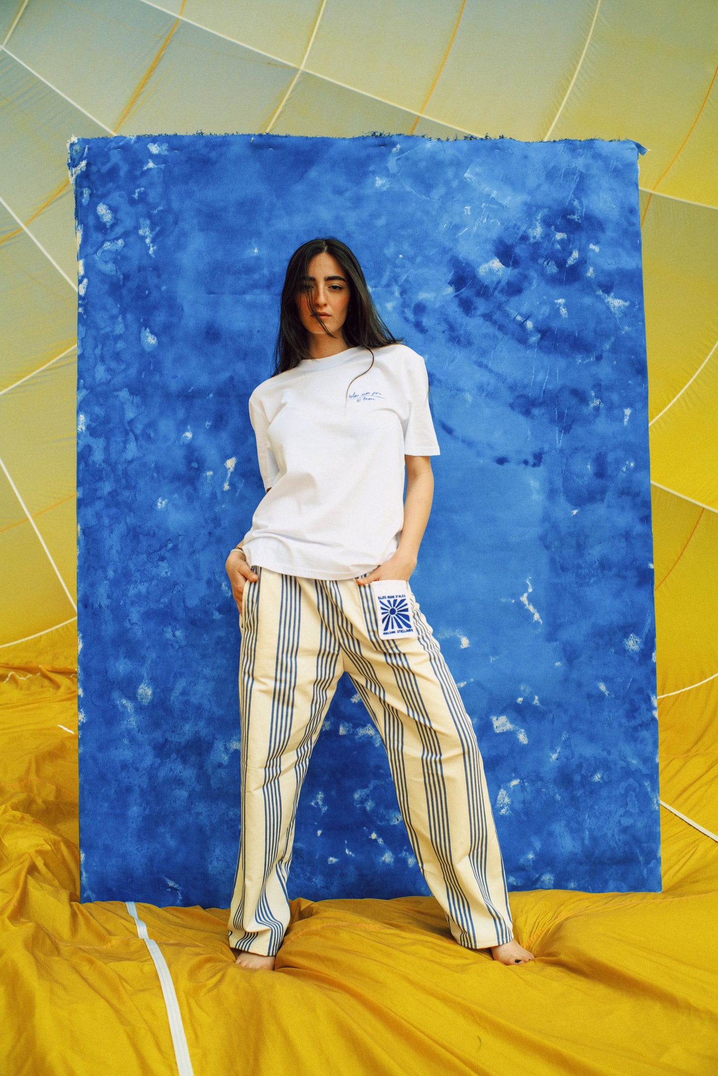 Femme posant devant un mur bleu et jaune – Style bohème chic, pantalon rayé et t-shirt blanc dans une atmosphère colorée et artistique.