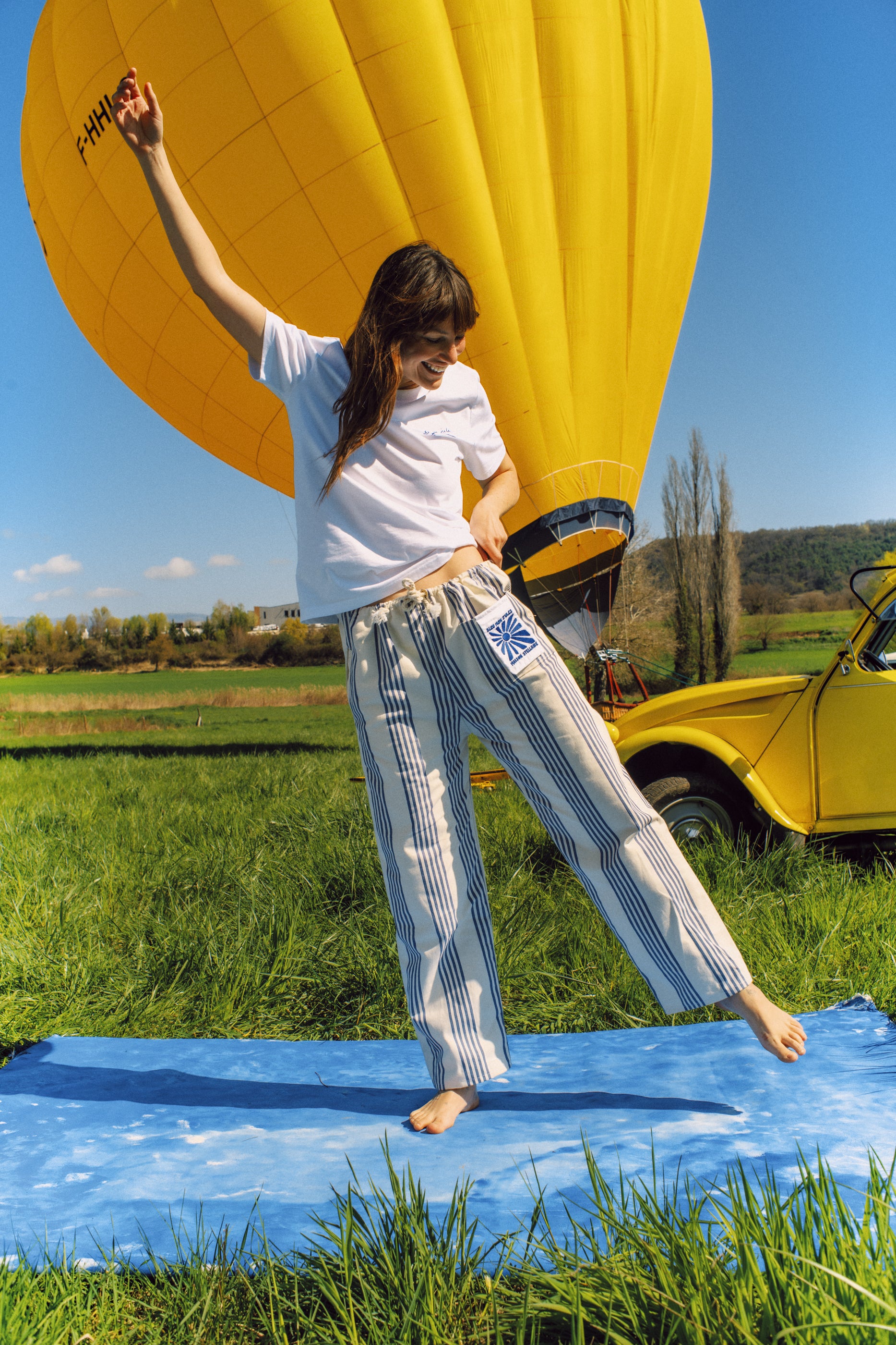 Femme portant le pantalon rayé dans un champ – Look d’été décontracté, pantalon rayé beige et bleu associé à un t-shirt blanc, devant une montgolfière jaune.