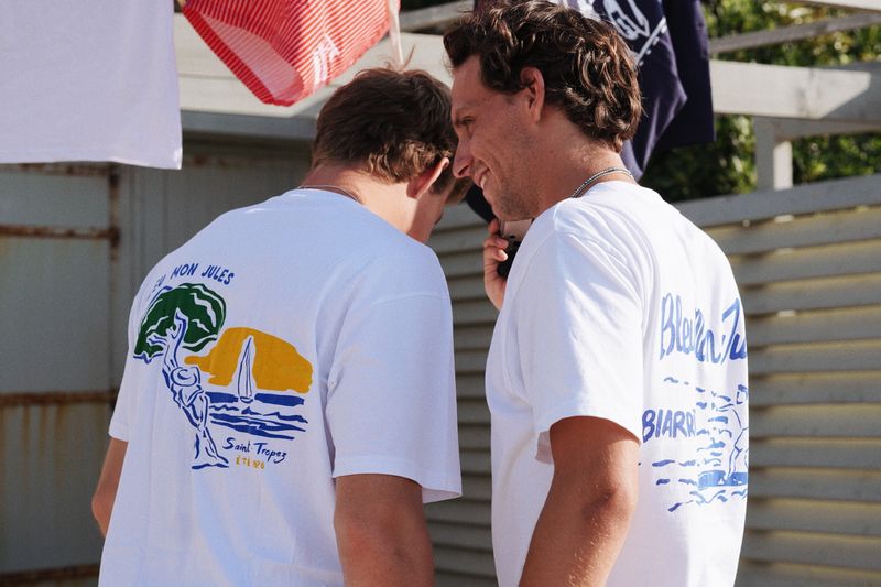Deux hommes portant des t-shirts blanc coton Saint-Tropez avec pin parasol et voilier bleu, été Côte d'Azur