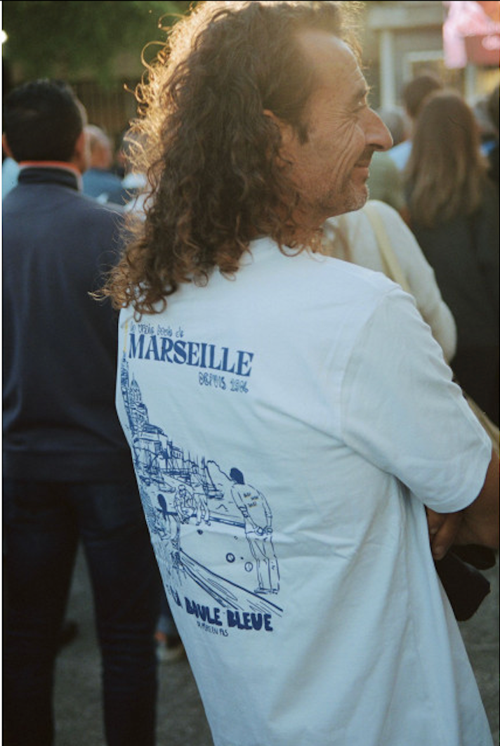 Homme portant t-shirt blanc Marseille La Boule Bleue avec illustration bateau, vue de profil en intérieur