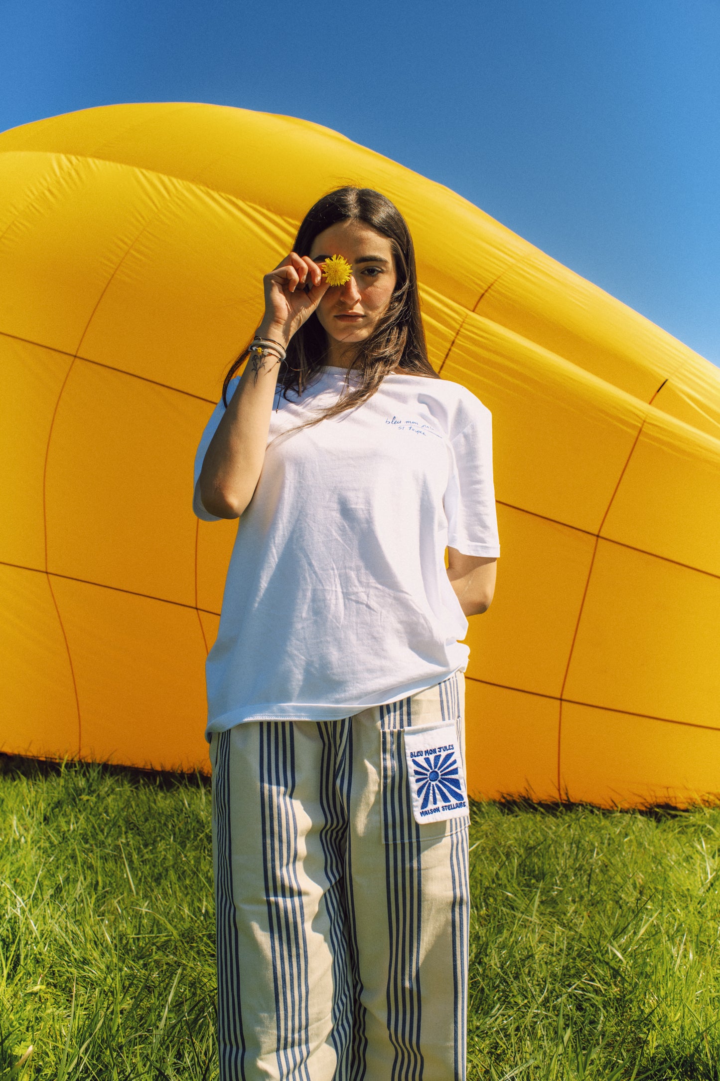 Femme debout devant une montgolfière – Tenue légère et estivale avec pantalon rayé bleu et beige, ambiance soleil et liberté.