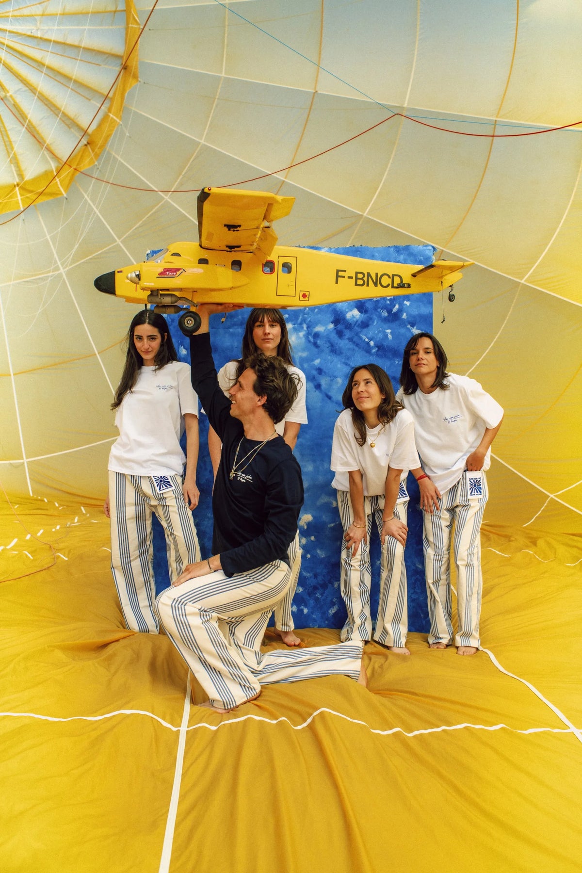 Cinq femmes en tenue blanc et bleu posent devant un avion jaune modèle réduit suspendu dans un décor de ballon géant