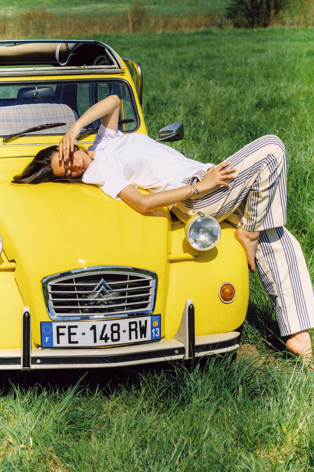 Femme allongée sur le capot d'une Citroën 2CV jaune vintage dans un champ verdoyant par beau temps