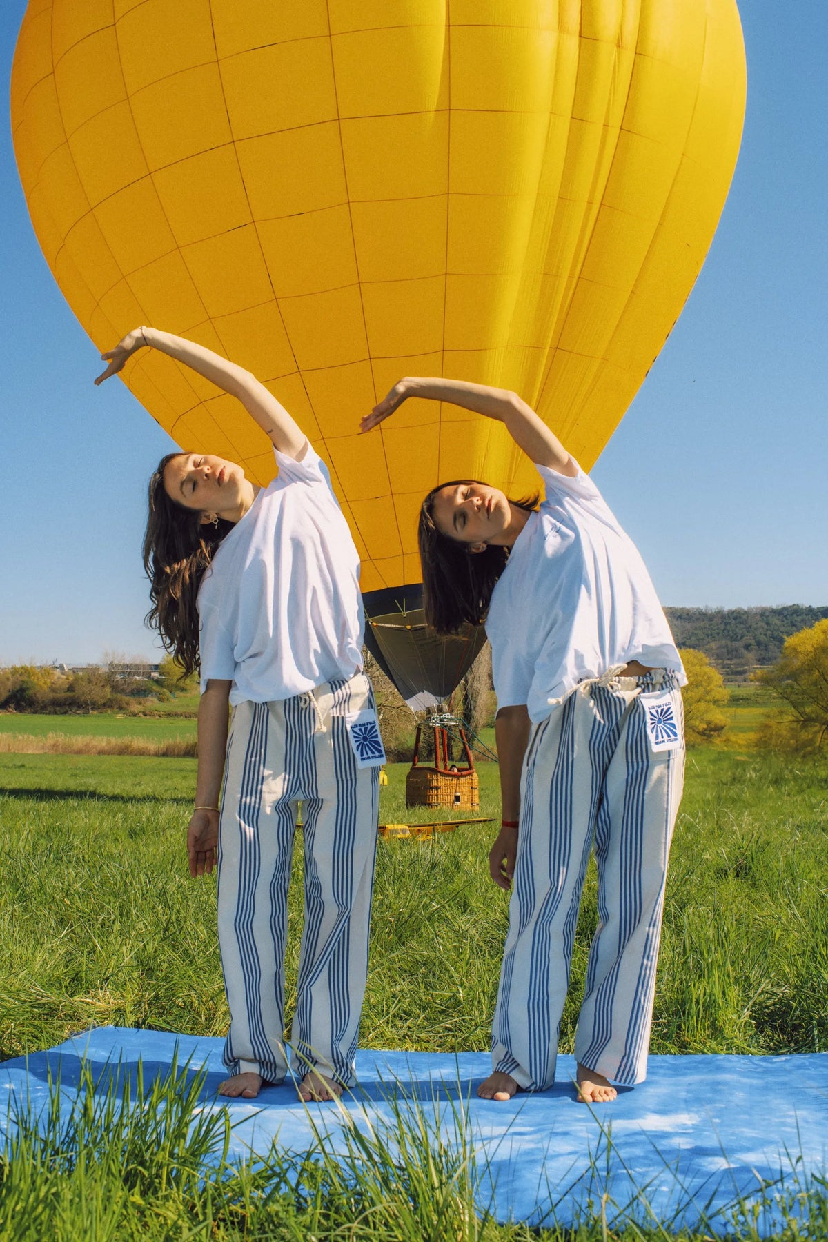 Deux femmes en tenue blanche et pantalon rayé bleu font un étirement sous un ballon jaune dans un champ