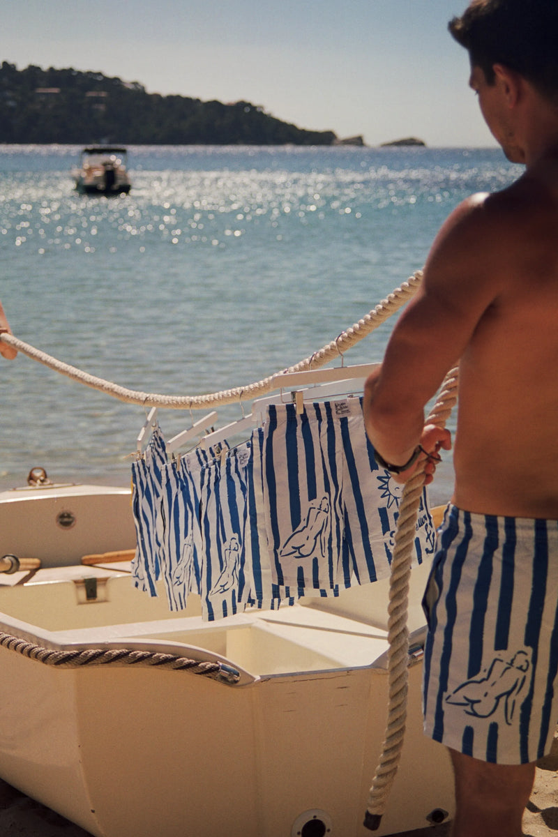 Homme sur un bateau à voile avec serviette rayée bleu blanc, mer turquoise et voilier en arrière-plan