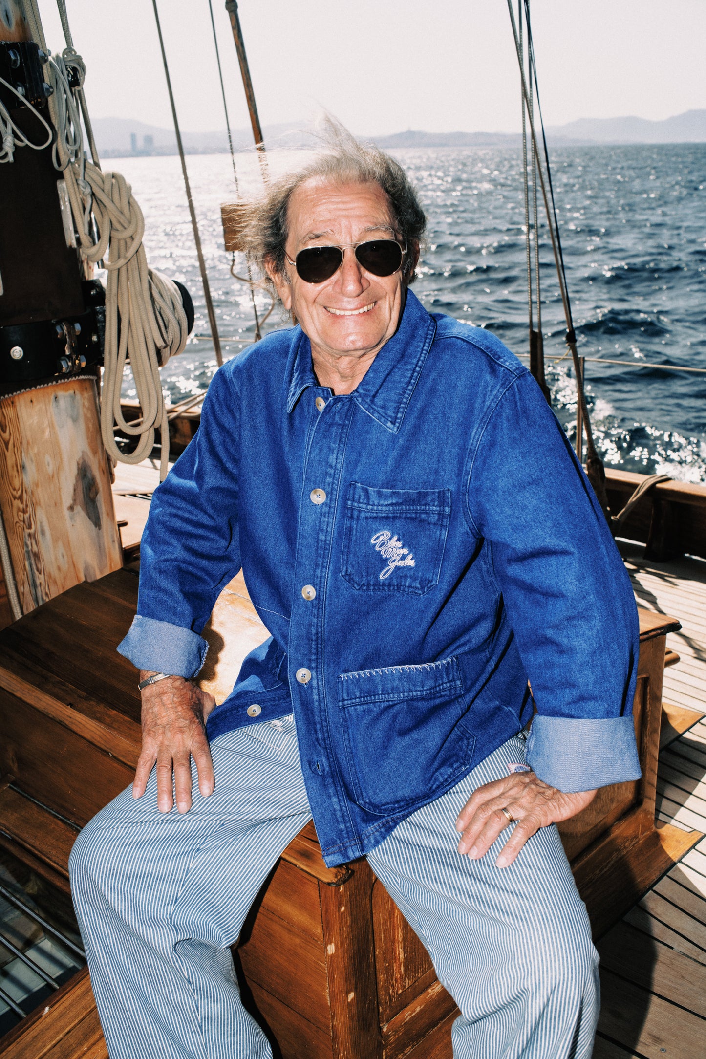 Homme âgé souriante en chemise denim bleue sur un voilier en mer, portant des lunettes de soleil aviateur