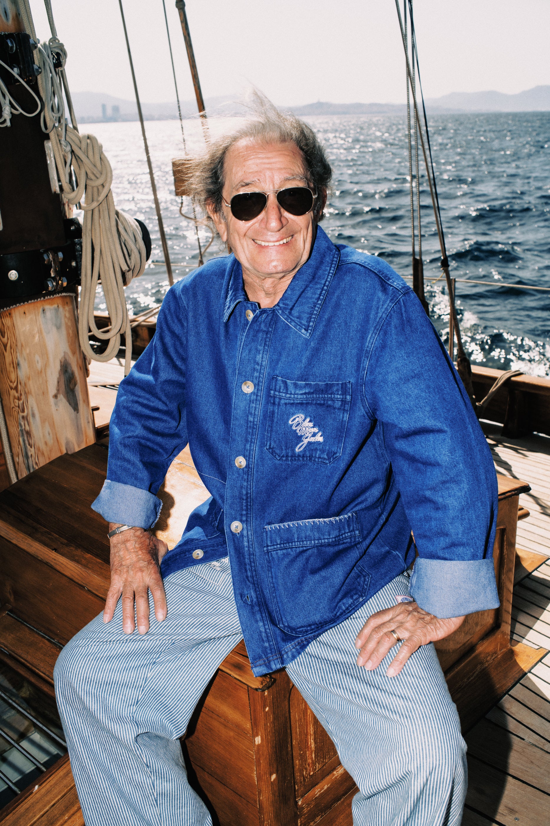 Homme âgé souriante en chemise denim bleue sur un voilier en mer, portant des lunettes de soleil aviateur