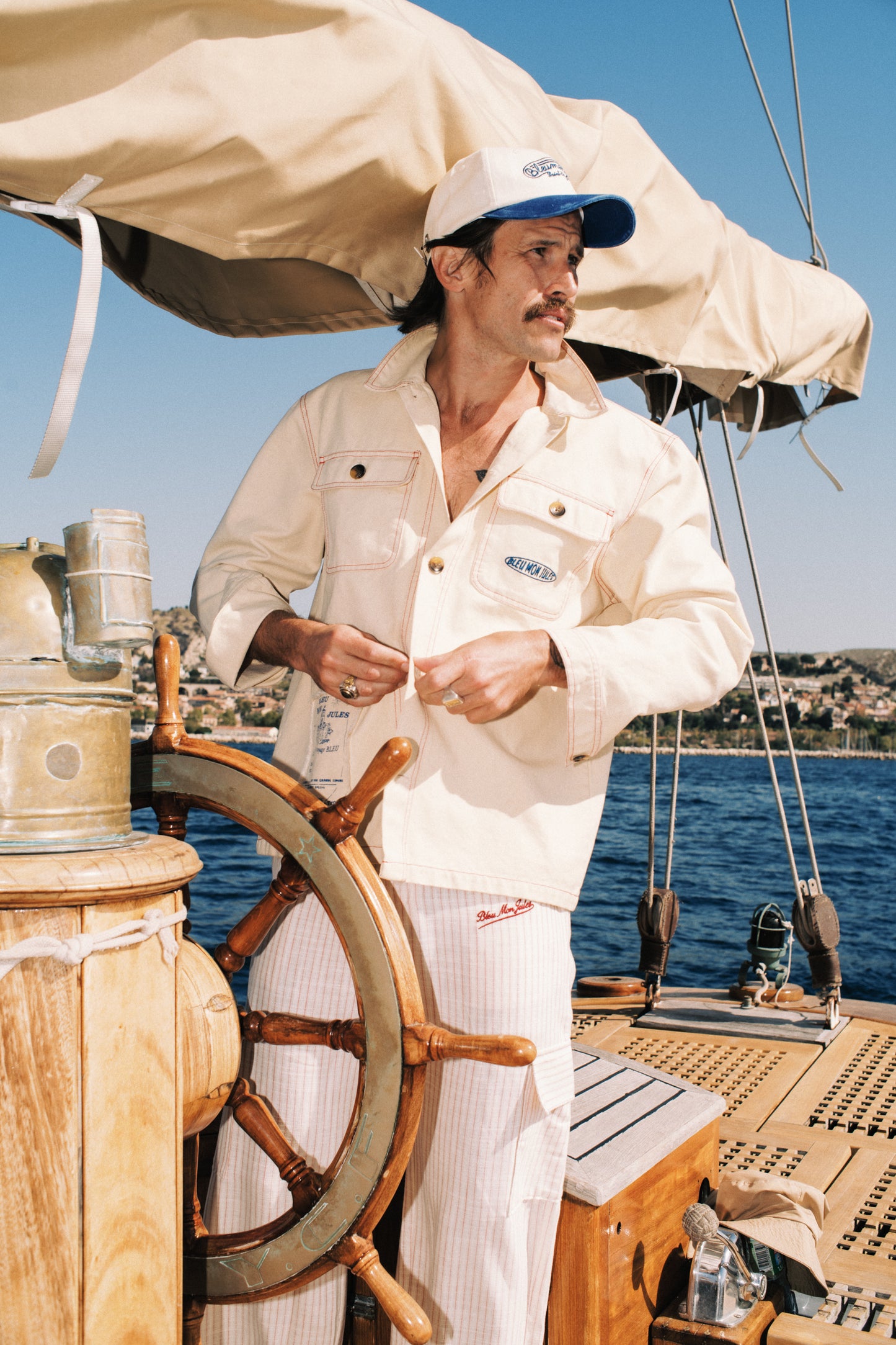 Homme à la barre d'un voilier portant la veste Bleu mon Jules crème, coton couleur sable, signature brodée poche, Saint-Tropez