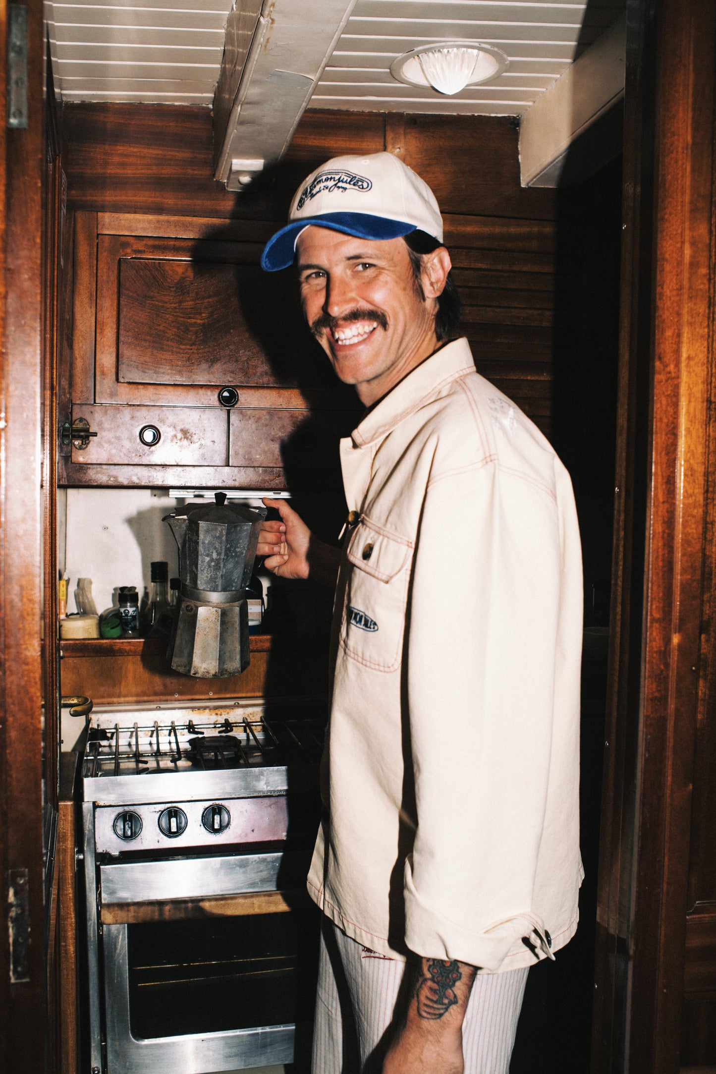 Homme portant veste crème coton Bleu mon Jules avec casquette, souriant en cuisine, style Saint-Tropez décontracté
