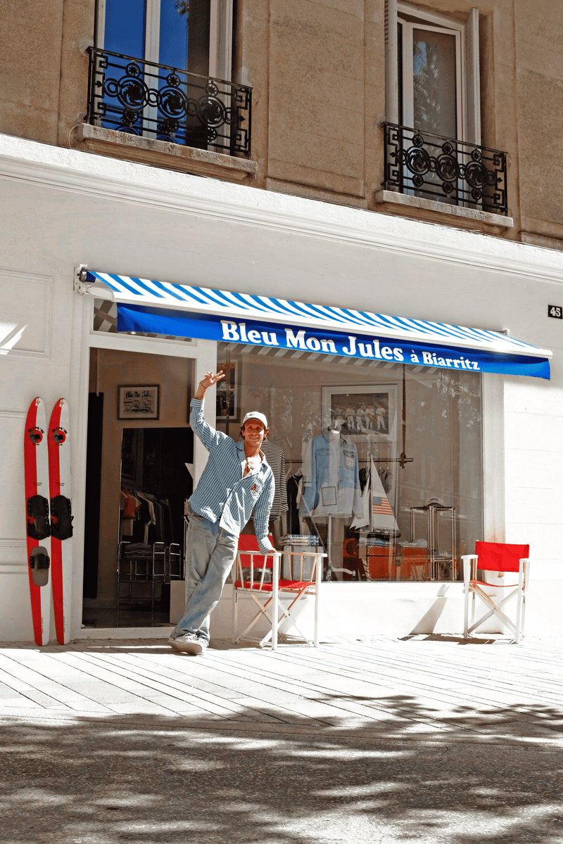 Homme posant devant boutique Bleu Mon Jules à Biarritz avec devanture bleue rayée, skis rouges et chaises blanches
