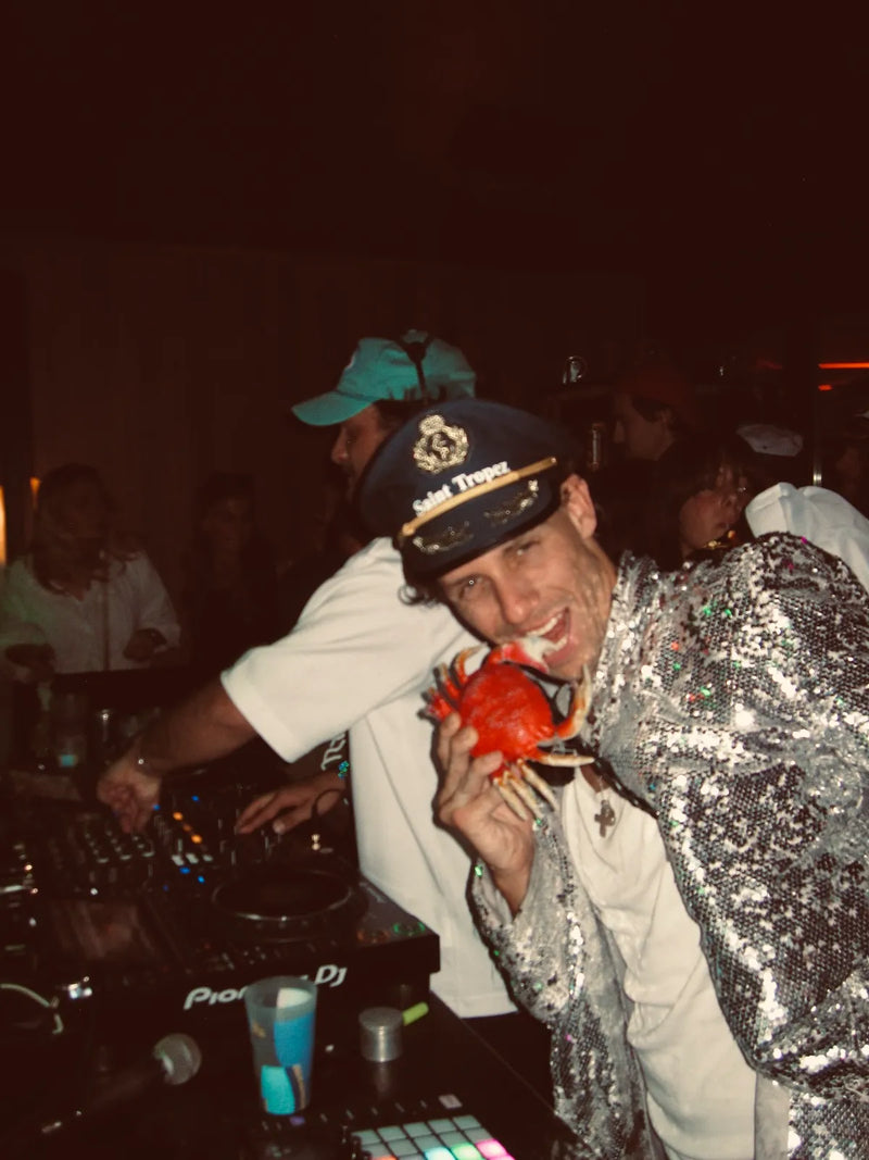 DJ animant une soirée en costume blanc et casquette bleue tenant un objet rouge devant public dansant